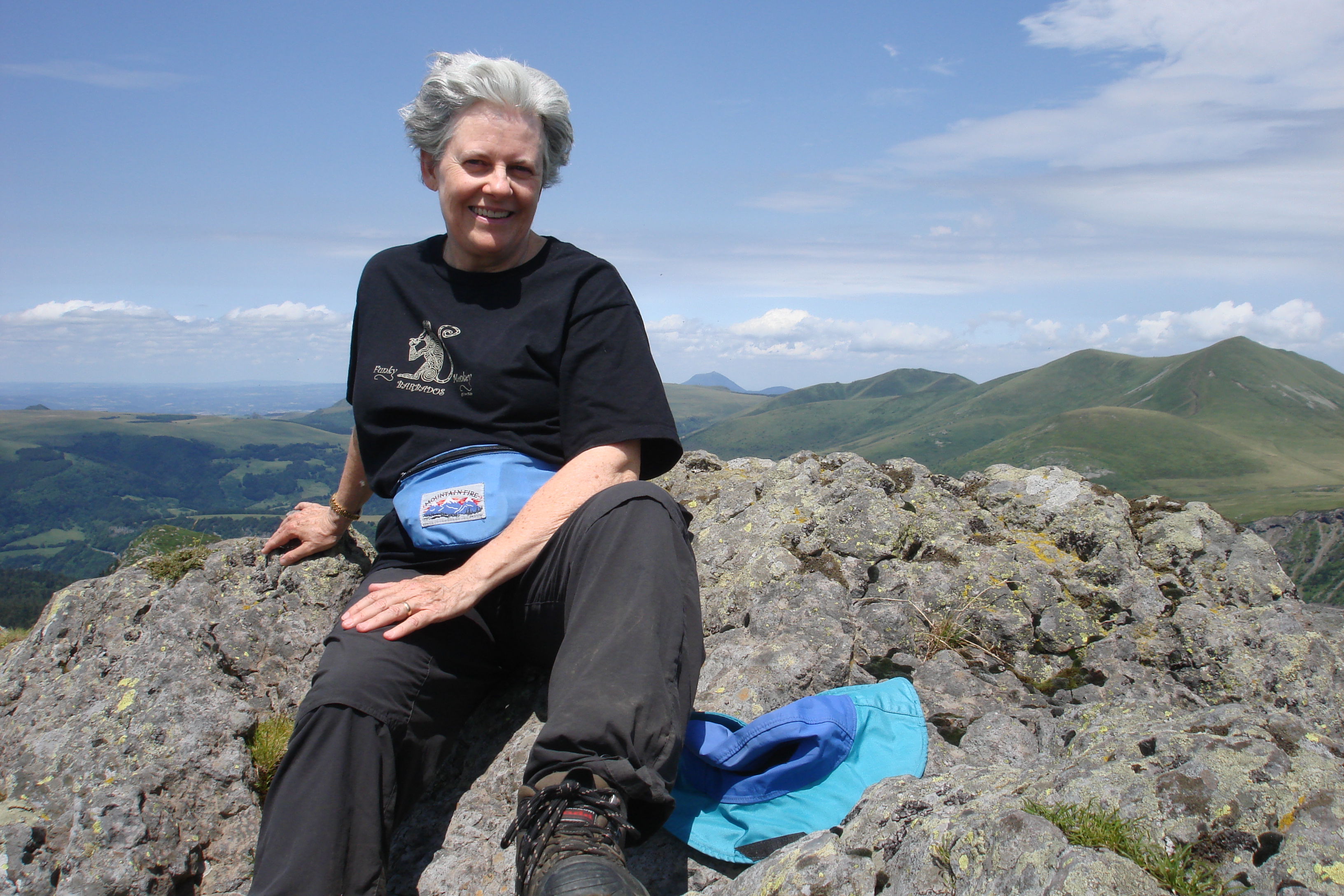 Claire wears athletic clothes and hiking boots. She is sitting on top of a mini volcano in the Massif-Centrale region of France overlooking a view of beautiful mountains and blue skies with some wispy clouds.