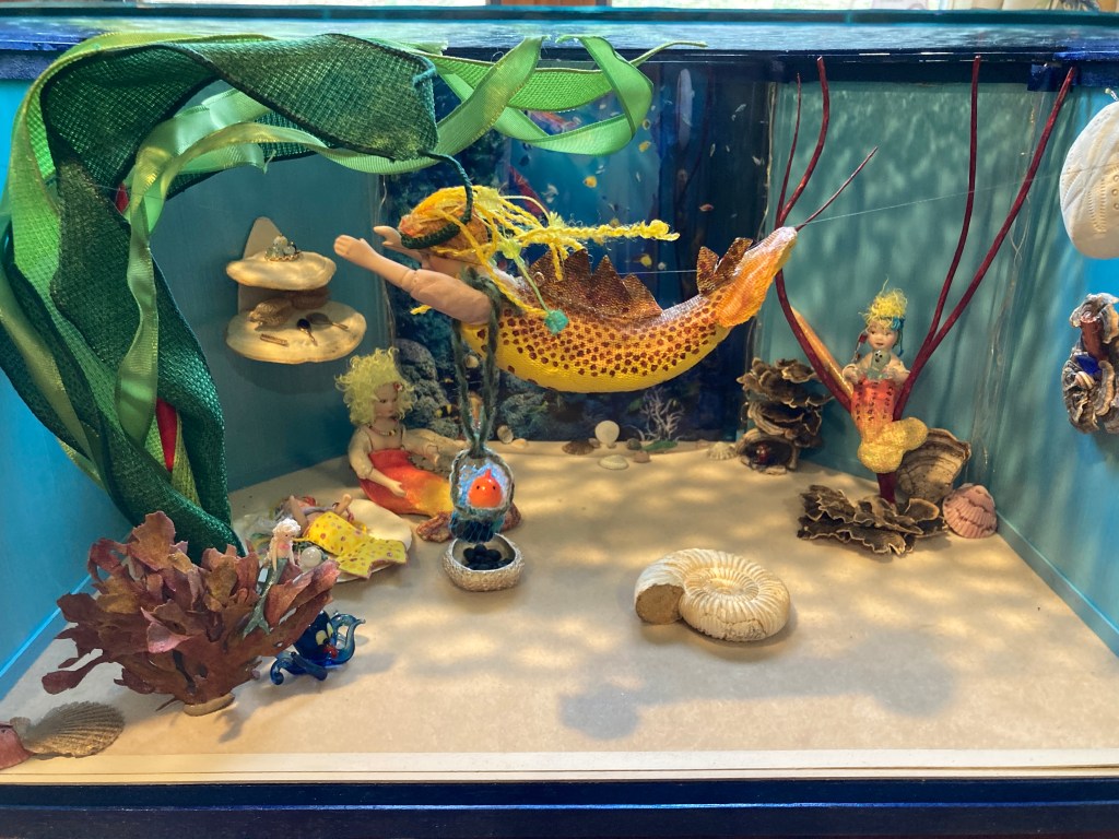 In this miniature diorama is a scene of mermaid family life underwater. Two mer-children play. The mom mermaid is swimming with long blonde hair and a yellow/orange spotted tail. There are different kinds of shells and seaweeds that make the miniature feel like an underwater room in a home.